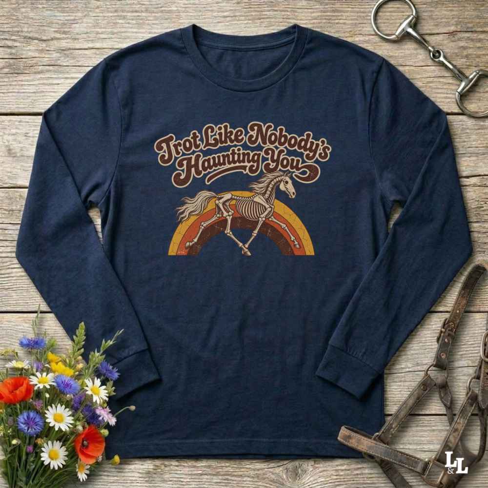 A navy blue long-sleeve t-shirt with a retro graphic of a skeleton horse running over a rainbow and the text 'Trot Like Nobody's Haunting You'. The shirt is displayed in a flat-lay arrangement on a rustic wooden background, surrounded by equestrian-themed props including a halter, a horse bit, and a small bouquet of wildflowers.