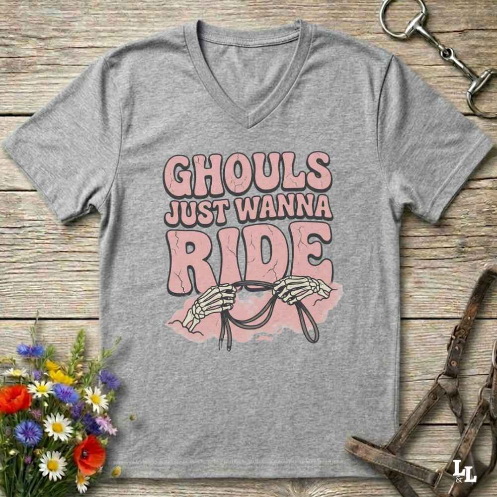 A heather gray V-neck t-shirt with a graphic print that reads 'GHOULS JUST WANNA RIDE' in pink and black, featuring skeleton hands holding reins over a pink splash. The shirt is displayed flat on a rustic wooden background with equestrian accessories and flowers.