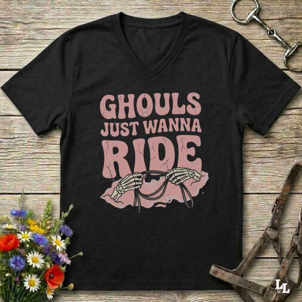 A black V-neck t-shirt with a pink graphic that reads 'GHOULS JUST WANNA RIDE' above skeleton hands holding reins, displayed on a rustic wooden background with equestrian accessories and flowers.