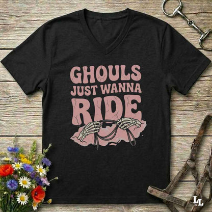 A black V-neck t-shirt with a pink graphic that reads 'GHOULS JUST WANNA RIDE' above skeleton hands holding reins, displayed on a rustic wooden background with equestrian accessories and flowers.