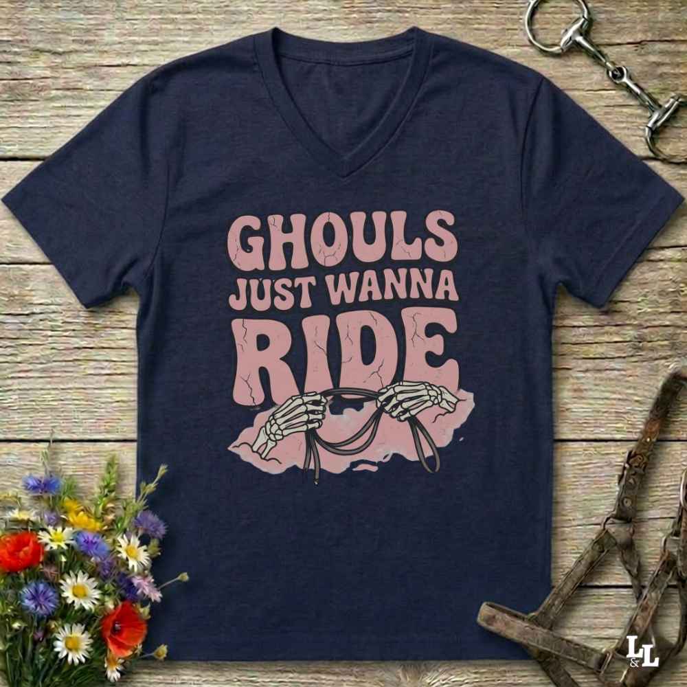 A navy blue V-neck t-shirt with a pink graphic that reads 'GHOULS JUST WANNA RIDE' above skeleton hands holding a horse bit, displayed on a rustic wooden background with horse tack and flowers.