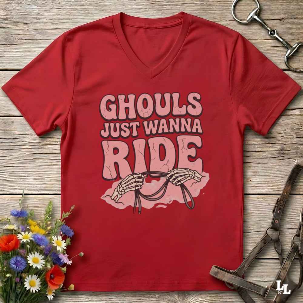 A red V-neck t-shirt with a graphic that reads 'GHOULS JUST WANNA RIDE' in pink and white, featuring skeleton hands holding reins, laid flat on a rustic wooden background with equestrian tack and flowers.