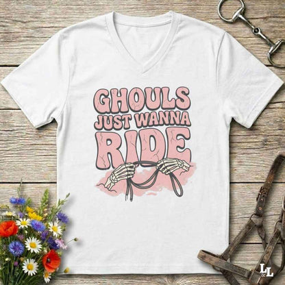 A white V-neck t-shirt with a graphic print that reads 'GHOULS JUST WANNA RIDE' in pink and brown lettering, featuring skeleton hands holding reins. The shirt is displayed flat on a rustic wooden background with equestrian tack and flowers as props.