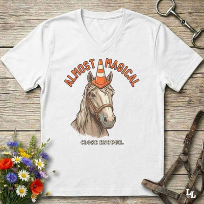 A white V-neck t-shirt laid flat on a rustic wooden surface, featuring a graphic of a horse wearing an orange and white traffic cone with the text 'ALMOST MAGICAL' above and 'CLOSE ENOUGH.' below.