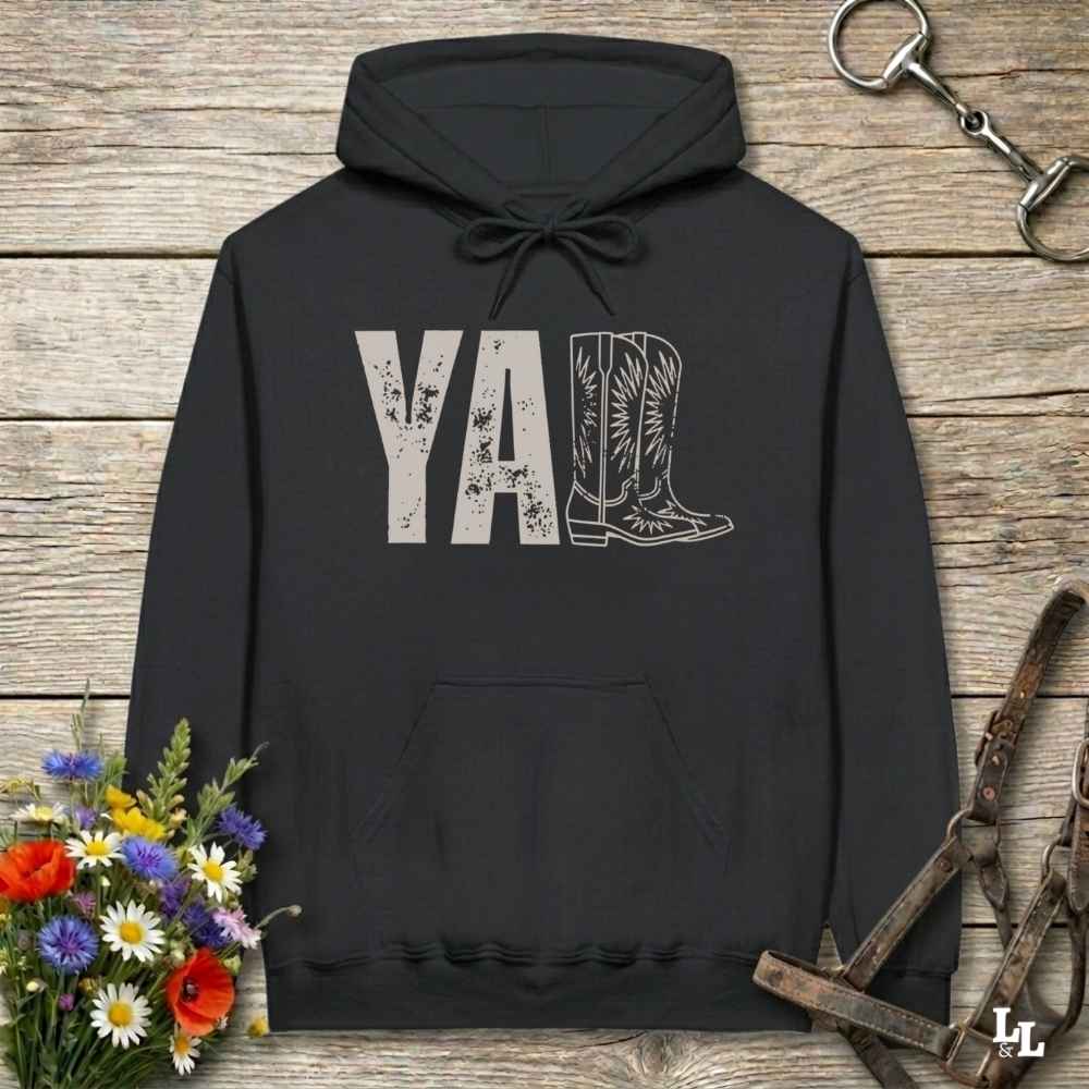 A black hoodie laid flat on a rustic wood plank surface, featuring a distressed 'Y'all' graphic with a cowboy boot illustration. The hoodie is styled with a bouquet of wildflowers, a horse bit, and a leather bridle around it.