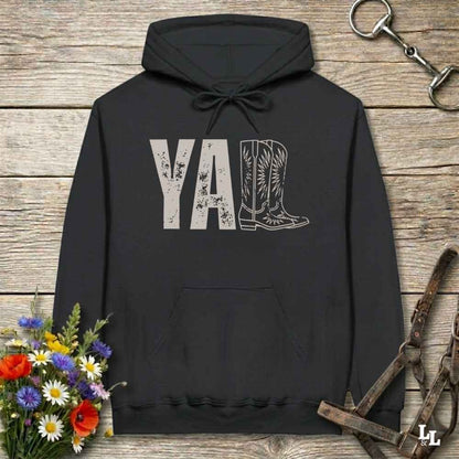 A black hoodie laid flat on a rustic wood plank surface, featuring a distressed 'Y'all' graphic with a cowboy boot illustration. The hoodie is styled with a bouquet of wildflowers, a horse bit, and a leather bridle around it.