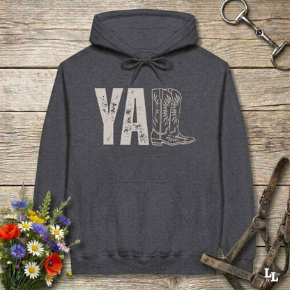 A dark heather gray hoodie laid flat on a rustic wood plank surface, featuring a distressed 'Y'all' graphic with a cowboy boot illustration. The hoodie is styled with a bouquet of wildflowers, a horse bit, and a leather bridle around it.