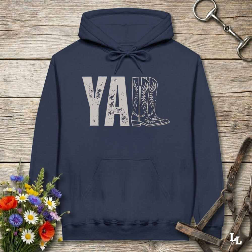 A navy blue hoodie laid flat on a rustic wood plank surface, featuring a distressed 'Y'all' graphic with a cowboy boot illustration. The hoodie is styled with a bouquet of wildflowers, a horse bit, and a leather bridle around it.