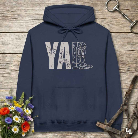 A navy blue hoodie laid flat on a rustic wood plank surface, featuring a distressed 'Y'all' graphic with a cowboy boot illustration. The hoodie is styled with a bouquet of wildflowers, a horse bit, and a leather bridle around it.