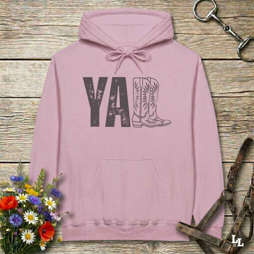 A pink hoodie laid flat on a rustic wood plank surface, featuring a distressed 'Y'all' graphic with a cowboy boot illustration. The hoodie is styled with a bouquet of wildflowers, a horse bit, and a leather bridle around it.