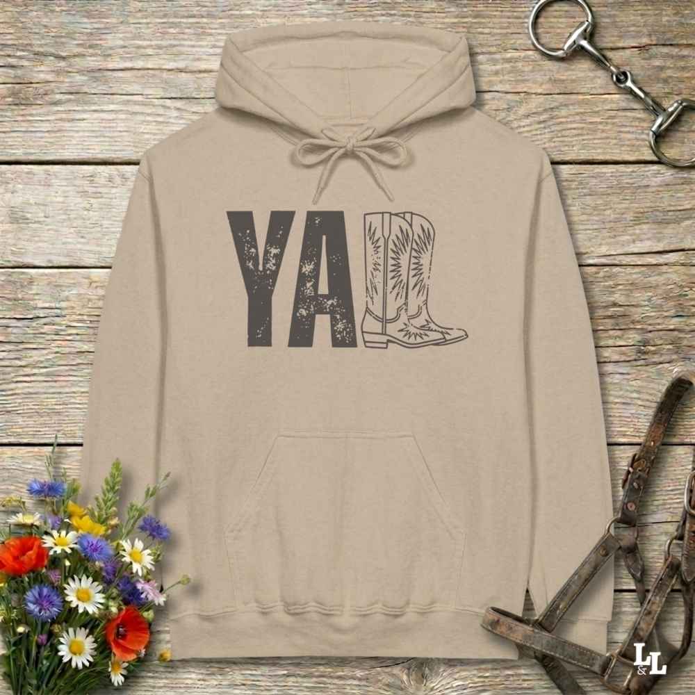 A beige hoodie laid flat on a rustic wood plank surface, featuring a distressed 'Y'all' graphic with a cowboy boot illustration. The hoodie is styled with a bouquet of wildflowers, a horse bit, and a leather bridle around it.