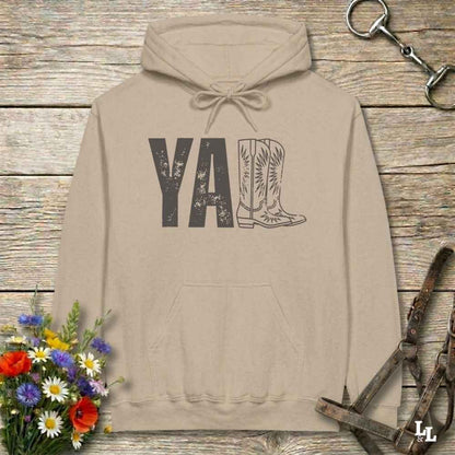 A beige hoodie laid flat on a rustic wood plank surface, featuring a distressed 'Y'all' graphic with a cowboy boot illustration. The hoodie is styled with a bouquet of wildflowers, a horse bit, and a leather bridle around it.