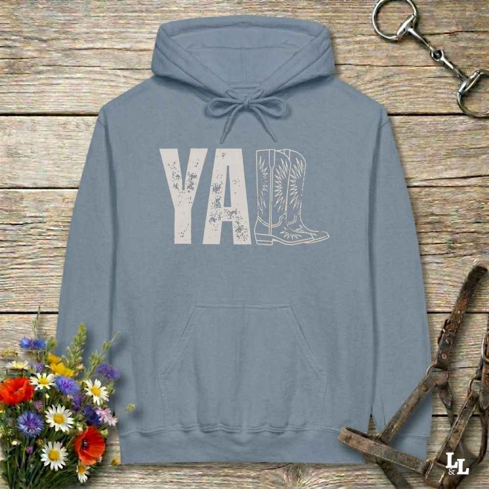 A light blue hoodie laid flat on a rustic wood plank surface, featuring a distressed 'Y'all' graphic with a cowboy boot illustration. The hoodie is styled with a bouquet of wildflowers, a horse bit, and a leather bridle around it.