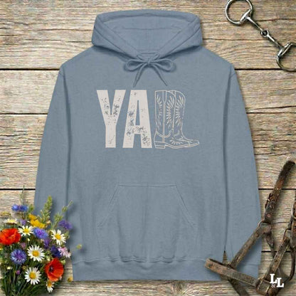 A light blue hoodie laid flat on a rustic wood plank surface, featuring a distressed 'Y'all' graphic with a cowboy boot illustration. The hoodie is styled with a bouquet of wildflowers, a horse bit, and a leather bridle around it.