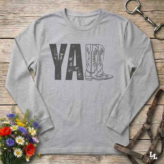 A light heather gray long-sleeve t-shirt with a graphic print reading 'Y'ALL' in distressed black letters, where the letters 'LL' are replaced by a western boot illustration. The shirt is laid flat on a rustic wooden surface with decorative props including a bouquet of flowers, a horse bit, and a leather bridle.