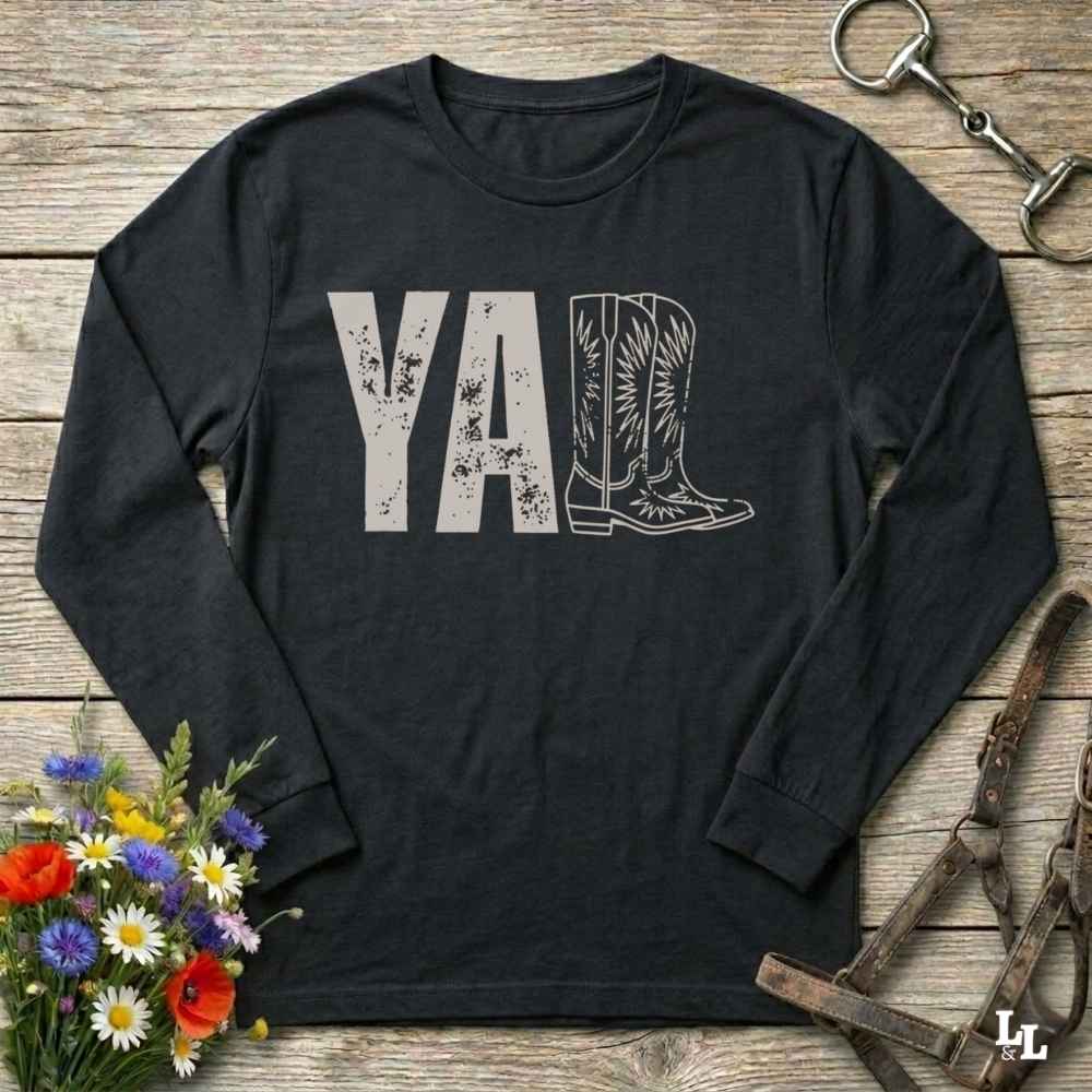 A black long-sleeve t-shirt with a graphic print reading 'Y'ALL' in distressed black letters, where the letters 'LL' are replaced by a western boot illustration. The shirt is laid flat on a rustic wooden surface with decorative props including a bouquet of flowers, a horse bit, and a leather bridle.