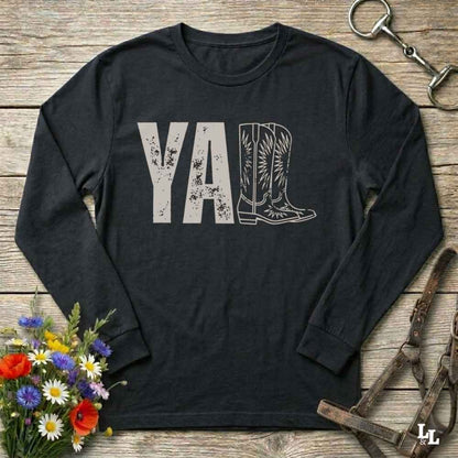 A black long-sleeve t-shirt with a graphic print reading 'Y'ALL' in distressed black letters, where the letters 'LL' are replaced by a western boot illustration. The shirt is laid flat on a rustic wooden surface with decorative props including a bouquet of flowers, a horse bit, and a leather bridle.