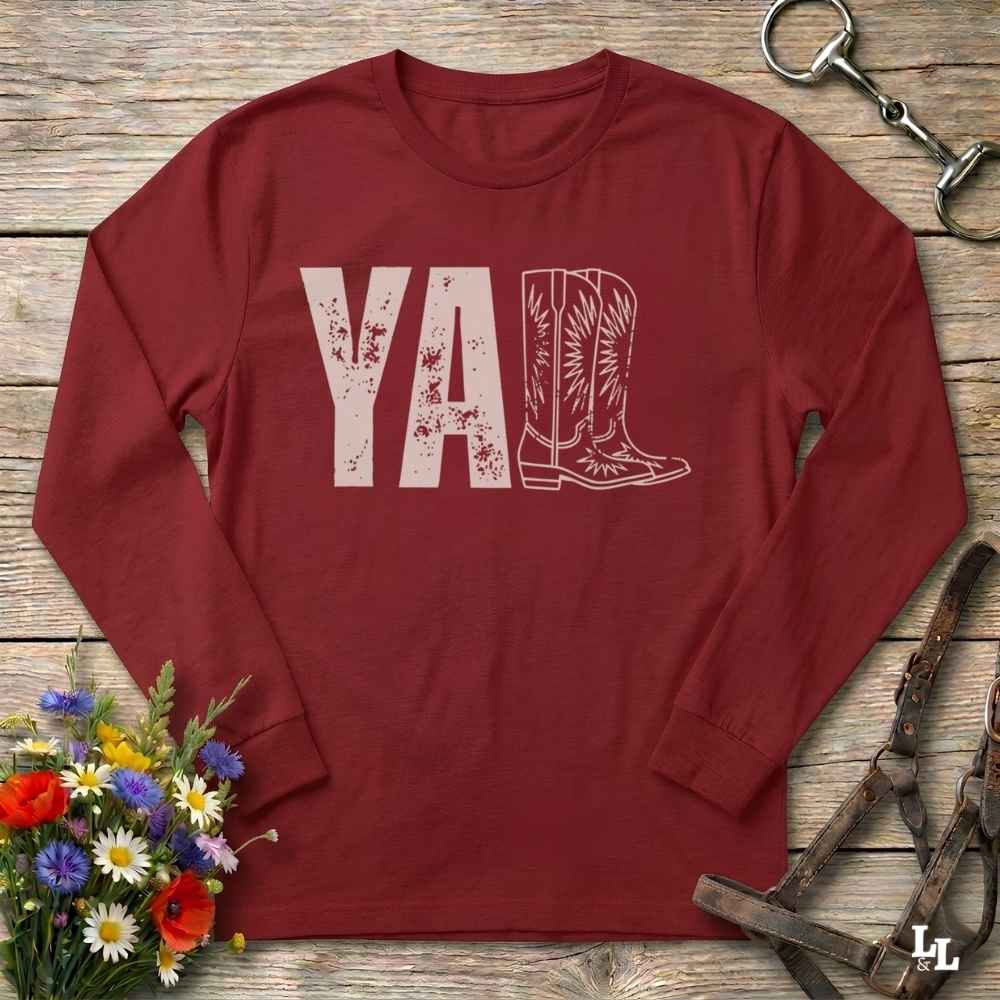 A red long-sleeve t-shirt with a graphic print reading 'Y'ALL' in distressed black letters, where the letters 'LL' are replaced by a western boot illustration. The shirt is laid flat on a rustic wooden surface with decorative props including a bouquet of flowers, a horse bit, and a leather bridle.