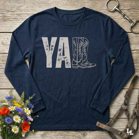 A navy blue long-sleeve t-shirt with a graphic print reading 'Y'ALL' in distressed black letters, where the letters 'LL' are replaced by a western boot illustration. The shirt is laid flat on a rustic wooden surface with decorative props including a bouquet of flowers, a horse bit, and a leather bridle.