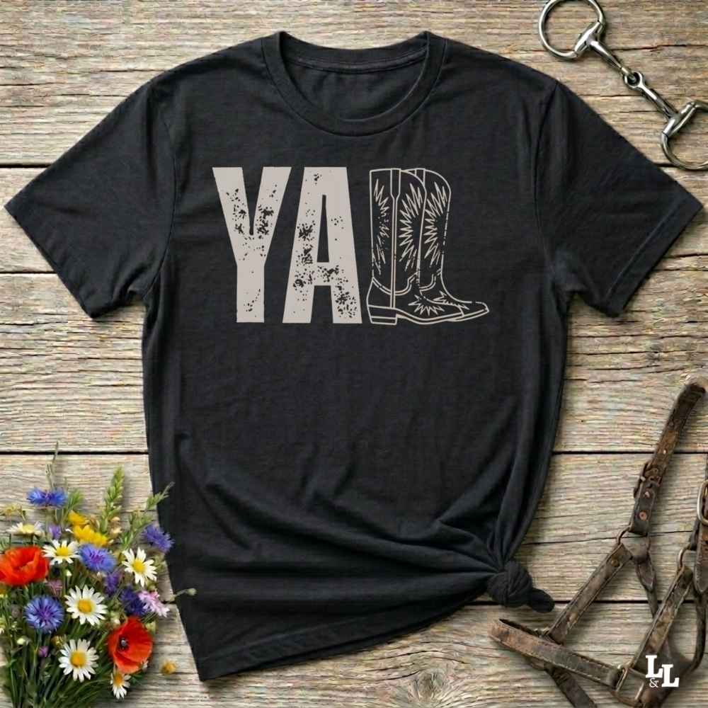 A black graphic t-shirt laid flat on a rustic wooden surface, styled with a small bouquet of wildflowers, a horse bit, and leather tack. The shirt features a dark gray graphic that reads 'YALL' next to a cowboy boot illustration.