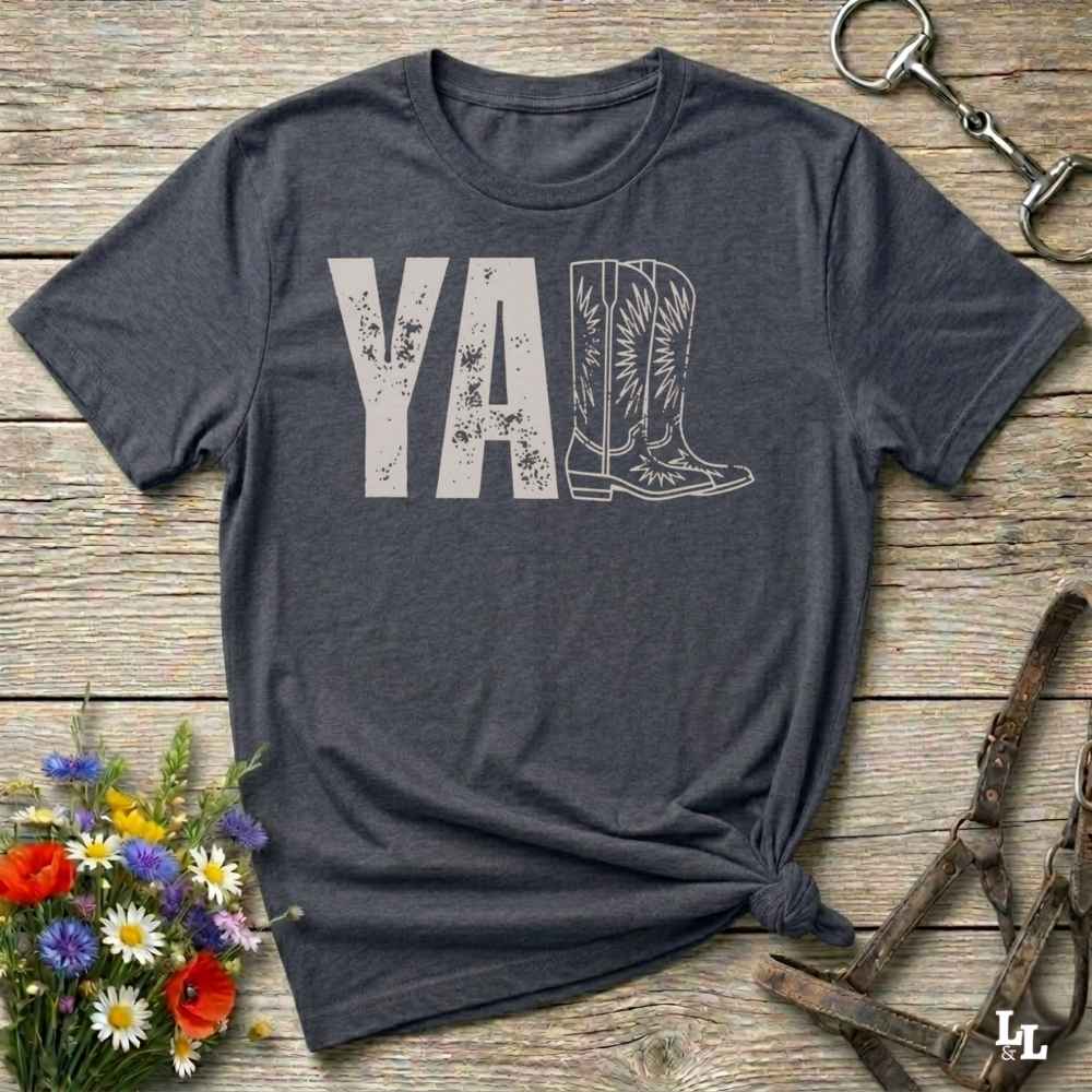 A dark grey graphic t-shirt laid flat on a rustic wooden surface, styled with a small bouquet of wildflowers, a horse bit, and leather tack. The shirt features a dark gray graphic that reads 'YALL' next to a cowboy boot illustration.