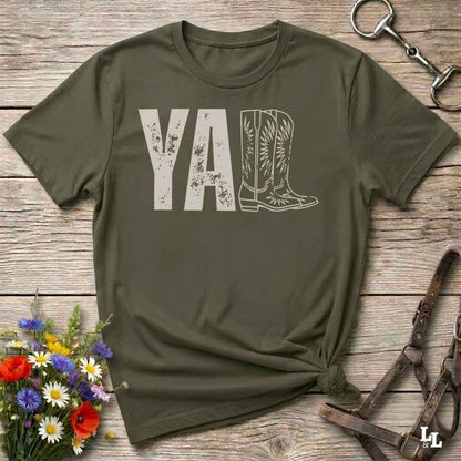 A green graphic t-shirt laid flat on a rustic wooden surface, styled with a small bouquet of wildflowers, a horse bit, and leather tack. The shirt features a dark gray graphic that reads 'YALL' next to a cowboy boot illustration.