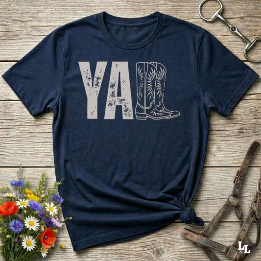 A navy blue graphic t-shirt laid flat on a rustic wooden surface, styled with a small bouquet of wildflowers, a horse bit, and leather tack. The shirt features a dark gray graphic that reads 'YALL' next to a cowboy boot illustration.