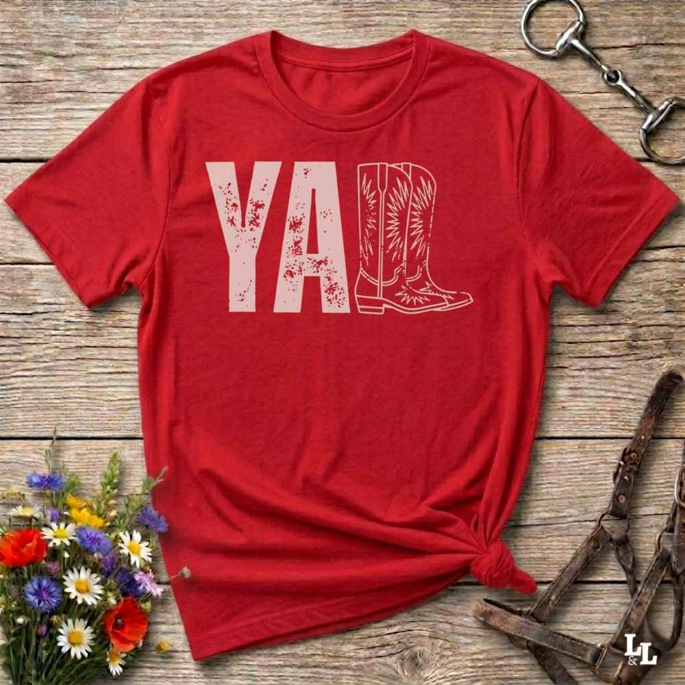 A red graphic t-shirt laid flat on a rustic wooden surface, styled with a small bouquet of wildflowers, a horse bit, and leather tack. The shirt features a dark gray graphic that reads 'YALL' next to a cowboy boot illustration.