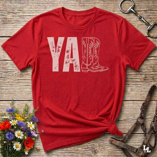 A red graphic t-shirt laid flat on a rustic wooden surface, styled with a small bouquet of wildflowers, a horse bit, and leather tack. The shirt features a dark gray graphic that reads 'YALL' next to a cowboy boot illustration.