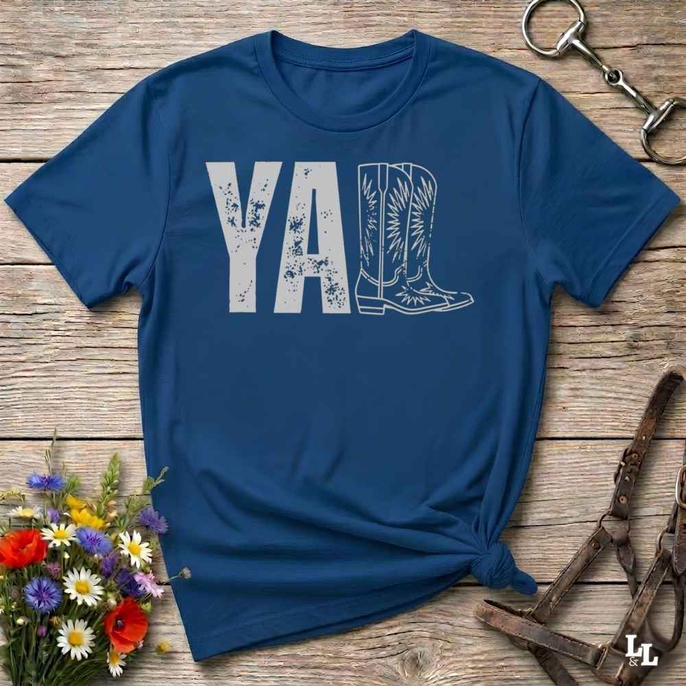 A blue graphic t-shirt laid flat on a rustic wooden surface, styled with a small bouquet of wildflowers, a horse bit, and leather tack. The shirt features a dark gray graphic that reads 'YALL' next to a cowboy boot illustration.