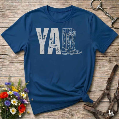A blue graphic t-shirt laid flat on a rustic wooden surface, styled with a small bouquet of wildflowers, a horse bit, and leather tack. The shirt features a dark gray graphic that reads 'YALL' next to a cowboy boot illustration.