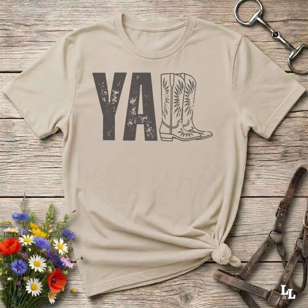 A beige graphic t-shirt laid flat on a rustic wooden surface, styled with a small bouquet of wildflowers, a horse bit, and leather tack. The shirt features a dark gray graphic that reads 'YALL' next to a cowboy boot illustration.