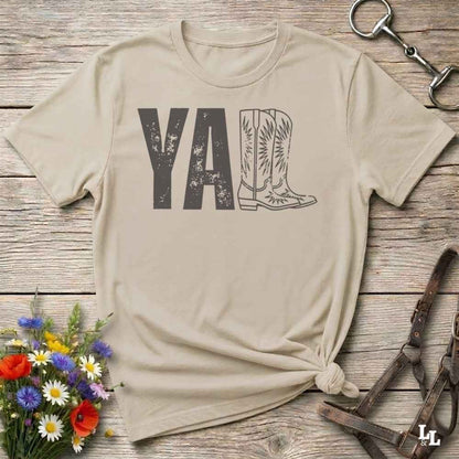 A beige graphic t-shirt laid flat on a rustic wooden surface, styled with a small bouquet of wildflowers, a horse bit, and leather tack. The shirt features a dark gray graphic that reads 'YALL' next to a cowboy boot illustration.