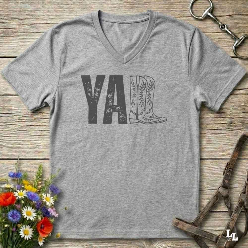 A heather gray V-neck t-shirt with a distressed graphic reading 'Y'ALL' where the 'll' is replaced by an illustration of cowboy boots, laid flat on a rustic wooden surface with equestrian and floral props.