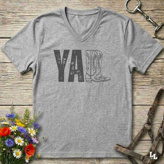 A heather gray V-neck t-shirt with a distressed graphic reading 'Y'ALL' where the 'll' is replaced by an illustration of cowboy boots, laid flat on a rustic wooden surface with equestrian and floral props.