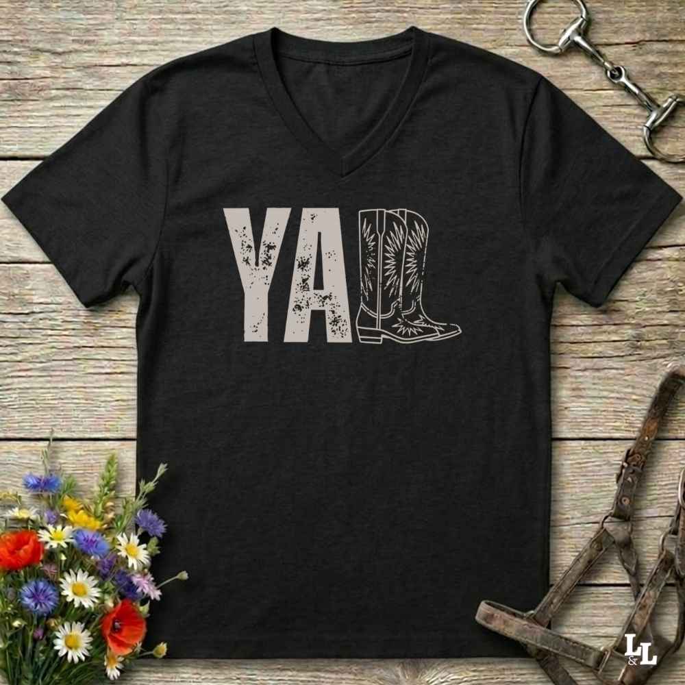 A black V-neck t-shirt with a distressed light gray graphic reading 'Y'ALL' where the letter 'l' is replaced by a cowboy boot illustration. The shirt is laid flat on a rustic wooden background with equestrian tack and wildflowers as props.