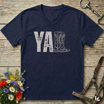 A navy blue V-neck t-shirt with a distressed white graphic reading 'Y'ALL' where the 'A' is replaced by a cowboy boot illustration. The shirt is laid flat on a rustic wooden background with equestrian-themed props like a horse bit, bridle, and a small bouquet of wildflowers.
