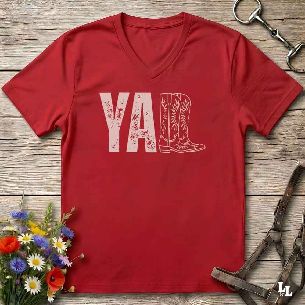 A red V-neck t-shirt with a distressed white graphic reading 'Y'ALL' and a cowboy boot illustration, laid flat on a rustic wood background with decorative flowers and horse tack.