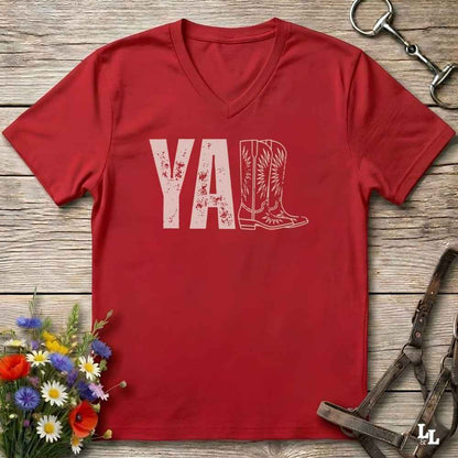 A red V-neck t-shirt with a distressed white graphic reading 'Y'ALL' and a cowboy boot illustration, laid flat on a rustic wood background with decorative flowers and horse tack.