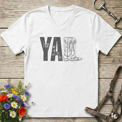 A white V-neck t-shirt with a western-themed graphic reading 'Y'ALL' in distressed black letters, where the letter 'L' is replaced by a cowboy boot illustration. The shirt is displayed flat on a rustic wooden background with equestrian tack and flowers.